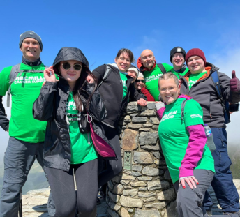 Nine PJH employees at the top of Snowden, with blue skies. The team wear green Macmillan tshirts along with warm clothing.